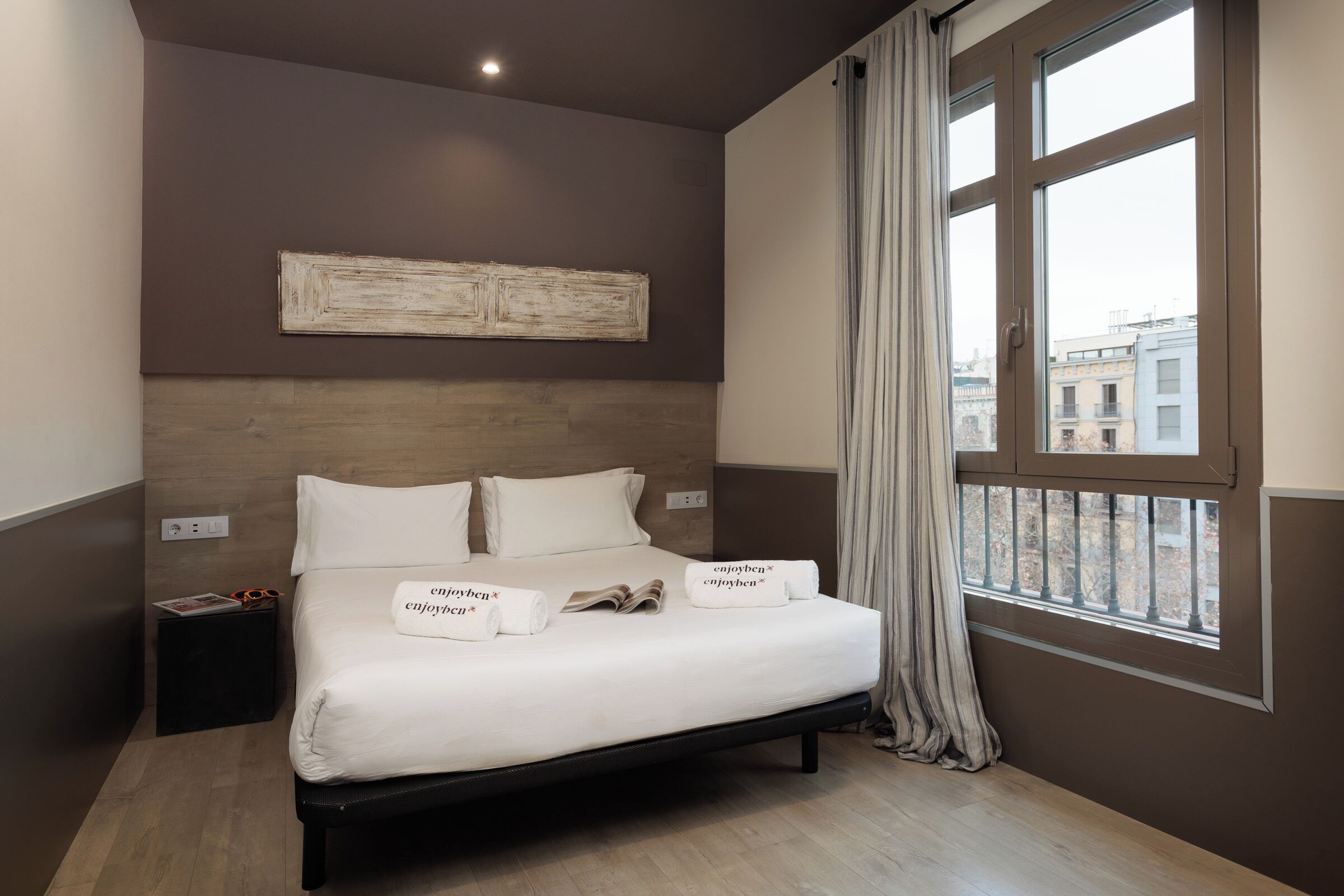 A neatly made bed with white linens and pillows, a bedside table with a magazine and glasses, and a view of buildings through a window with curtains.