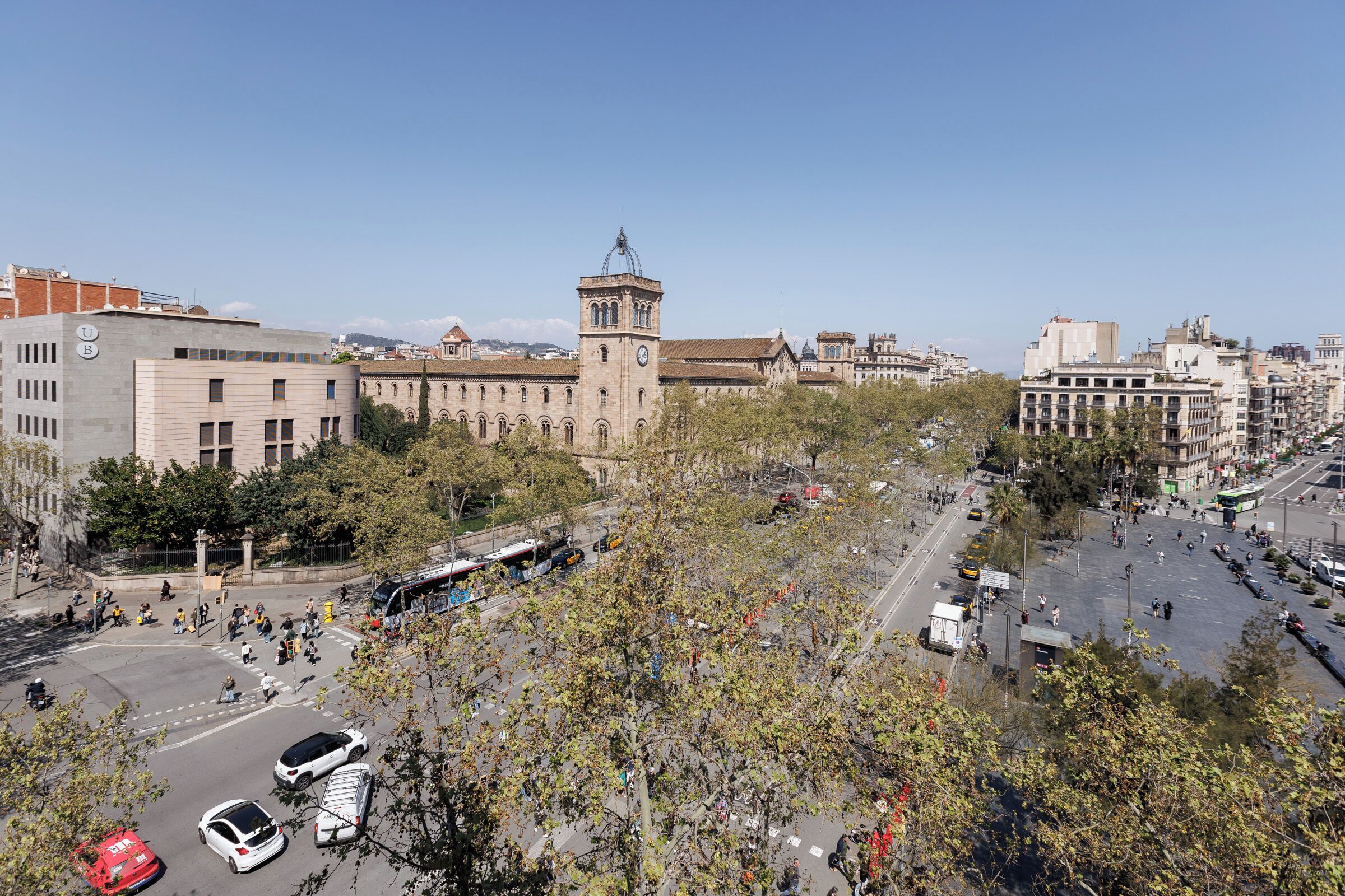 Un edifici gran amb una torre de l'ajuntament, envoltat d'arbres i un carrer animat amb cotxes i autobusos.