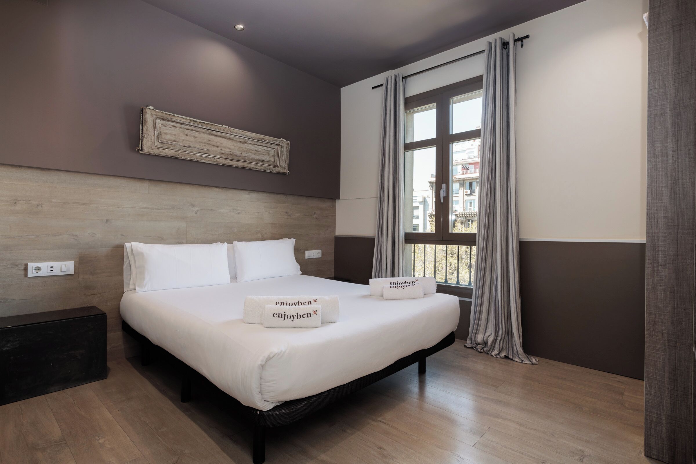 Een moderne slaapkamer met een groot bed, een nachtkastje, een houten hoofdbord en een raam met gordijnen.