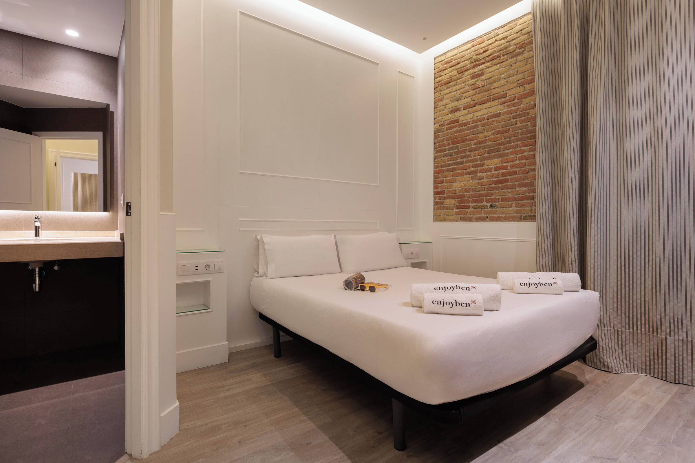 Een moderne hotelkamer met een groot bed, een houten vloer, een accentmuur van baksteen en een badkamer met spiegel en wastafel.