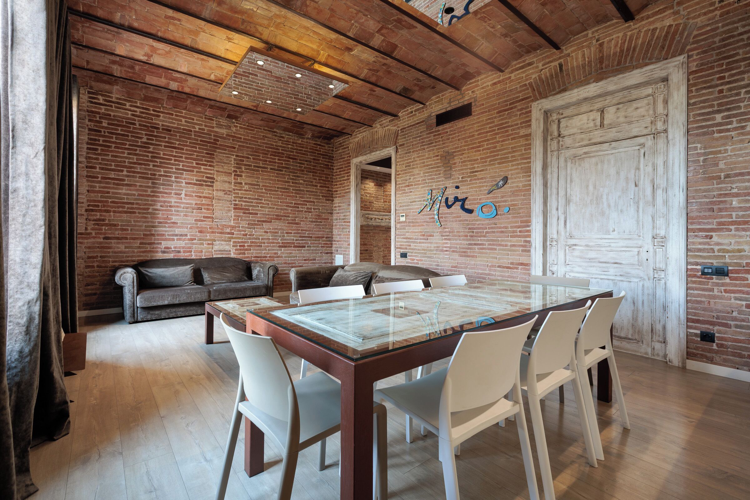 Un coin repas avec une table à plateau en verre et des chaises blanches, un mur en briques et un plafond en bois.