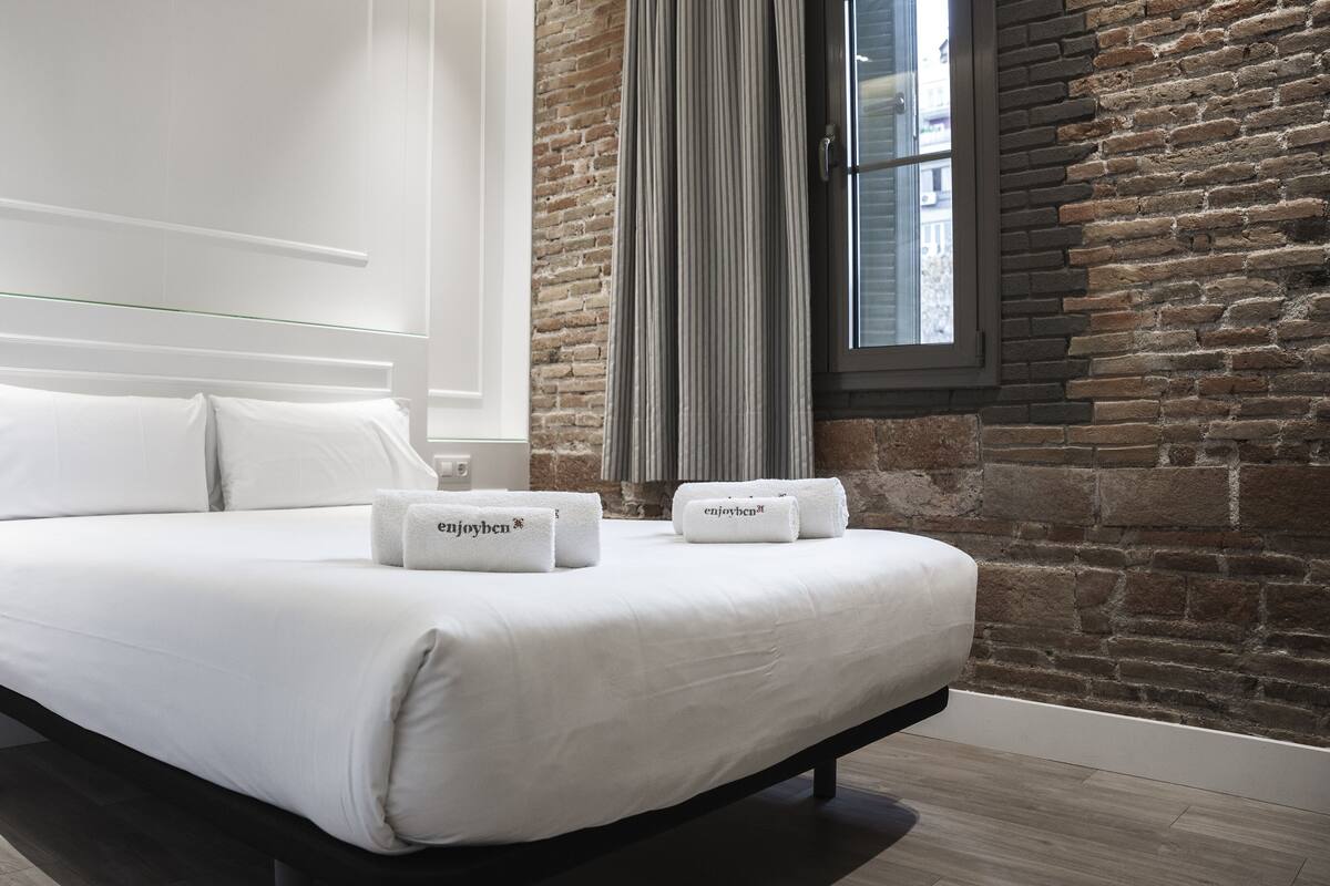 A modern bedroom with a large bed, white linens, and two towels with "enjoyben" branding.
