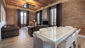 A spacious room with a large glass-top table, white chairs, a brown sofa, and a brick wall. - Enjoybcn Miro Apartments (Barcelona)