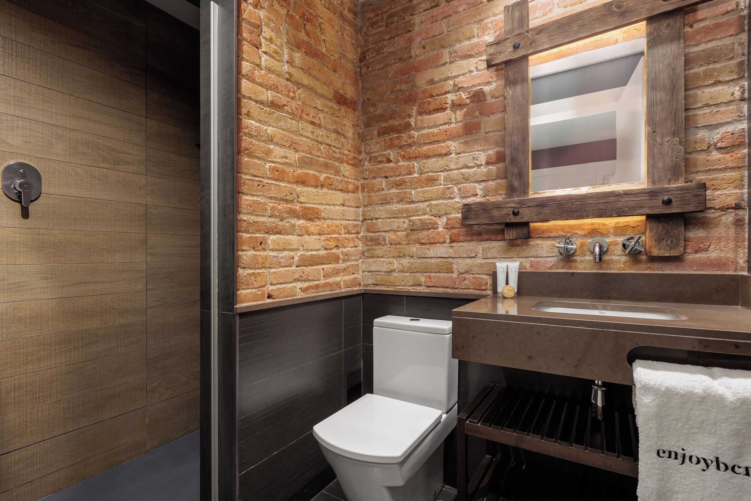 A modern bathroom with a brick wall, a wooden door, a white toilet, a sink with a mirror, and a towel hanging on a rack.