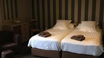 A double bed with two pillows and a blanket, a chair, a small table, and a lamp in a room with striped walls.