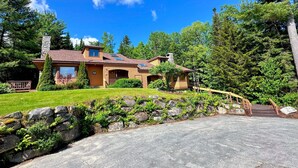 A two-story house with a stone chimney, a wooden deck, and a stone wall with greenery.