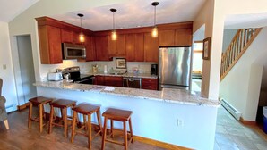 A kitchen with wooden cabinets, a stainless steel refrigerator, a microwave, and a granite countertop. There are three wooden stools at the island.