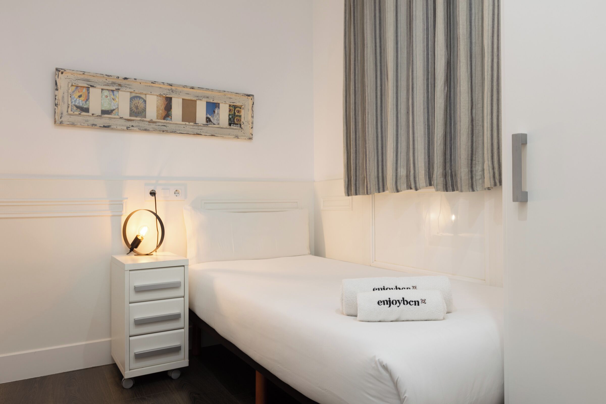 Une chambre d’hôtel bien rangée, avec un lit, une table de chevet dotée d’une lampe, une œuvre d’art accrochée au mur et une fenêtre avec des rideaux.
