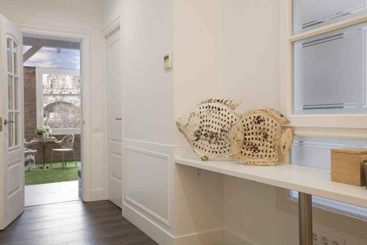 Un couloir avec une porte blanche donnant sur un balcon, une étagère blanche ornée d’objets décoratifs et une fenêtre avec des stores.