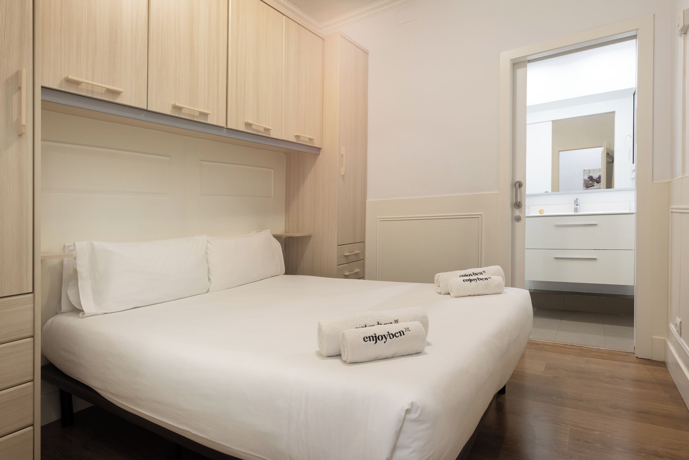 Una cama bien hecha con sábanas blancas y dos toallas con texto, un armario de madera, y un baño blanco con espejo y lavamanos.
