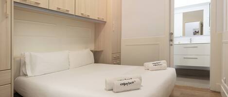 Una cama bien hecha con sábanas blancas y dos toallas con texto, un armario de madera, y un baño blanco con espejo y lavamanos.