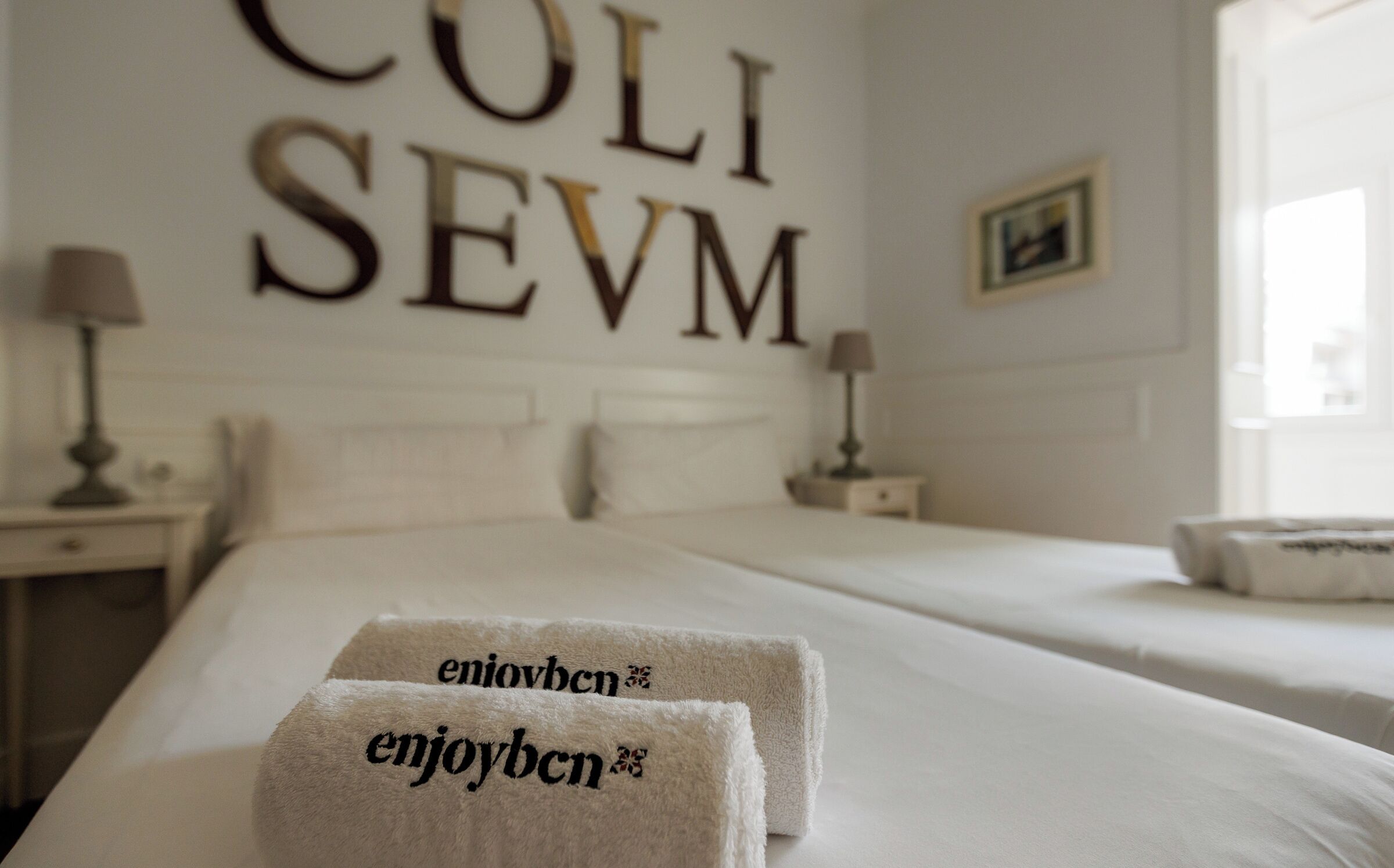 Une chambre d’hôtel avec un grand lit, deux oreillers, une table de chevet avec une lampe et un mur sur lequel est inscrit le texte « COLI SEVM ».