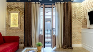 A room with a red sofa, a glass coffee table with a magazine, and a potted plant. There is a large window with curtains, a brick wall, and a door leading outside. - Enjoybcn Colon Apartments (Barcelona)