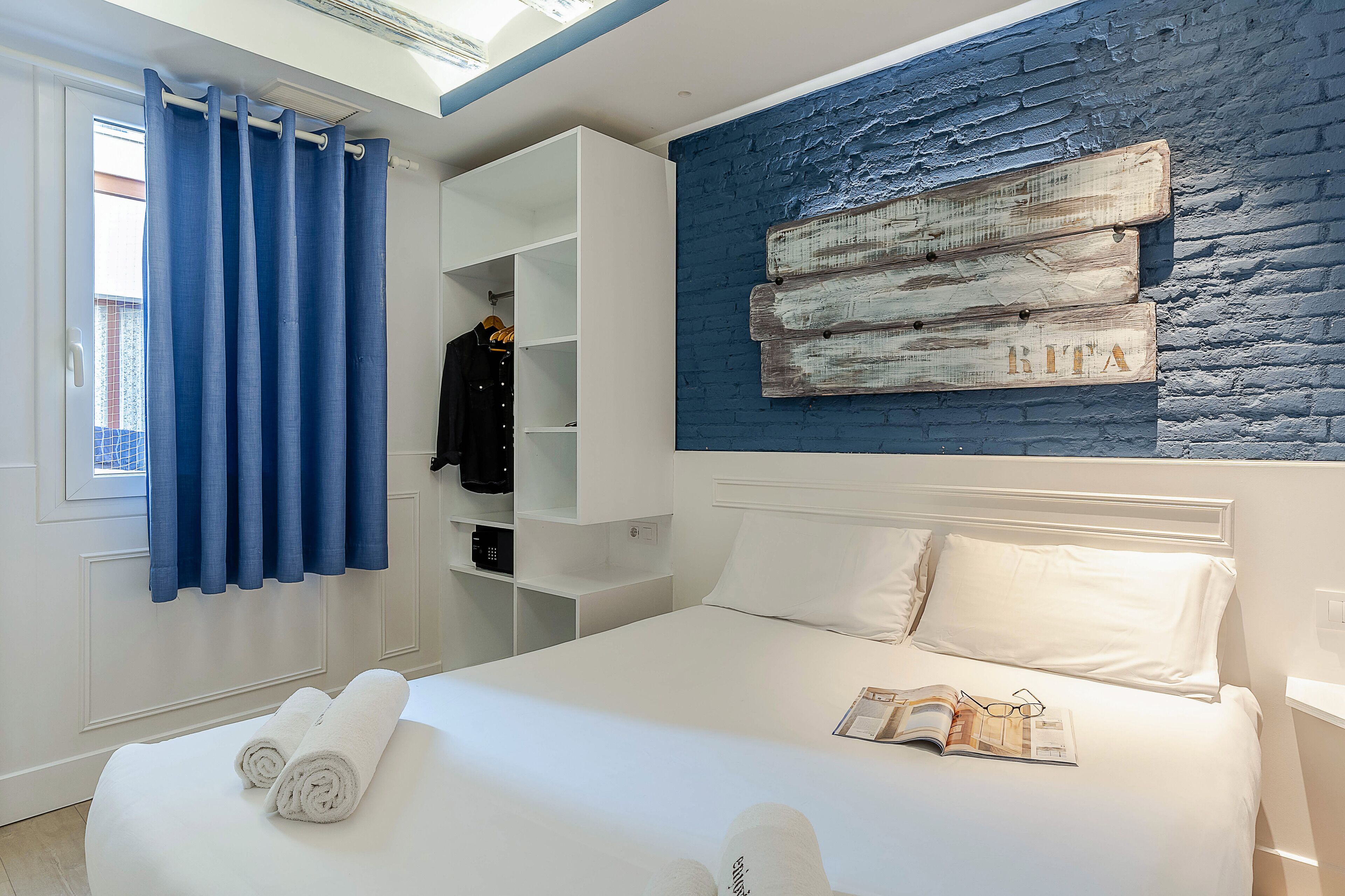 Een slaapkamer met een bed, een raam met blauwe gordijnen, een witte kledingkast en een blauwe bakstenen muur met een houten bord.