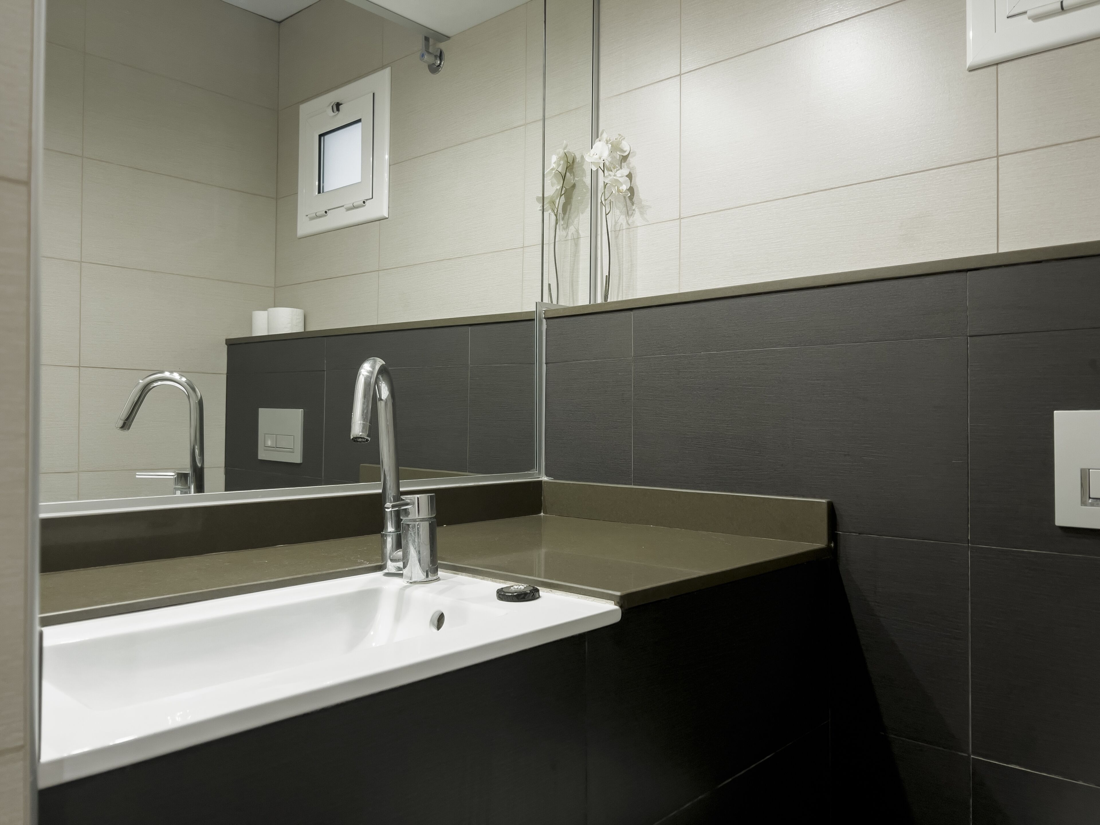 A modern bathroom with a white sink, a chrome faucet, and dark tiled walls.