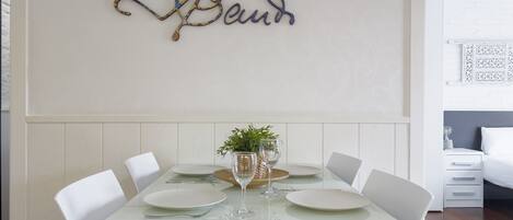 A dining area with a white table set for four, surrounded by white chairs. A decorative wall piece spelling "Baun" hangs above the table.