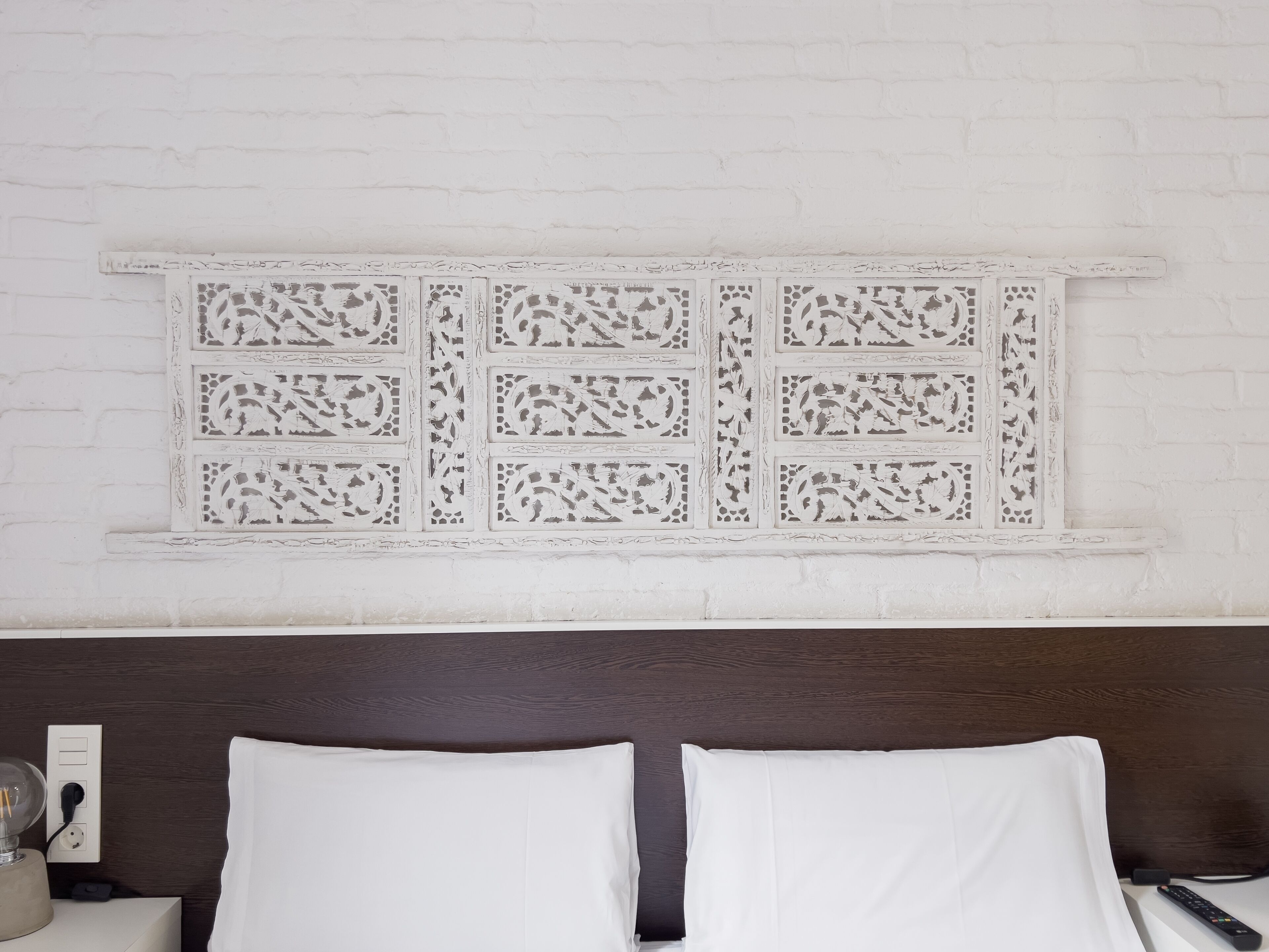Un dormitorio con pared de ladrillo blanco, un panel decorativo blanco sobre la cama y cabecera de madera oscura.