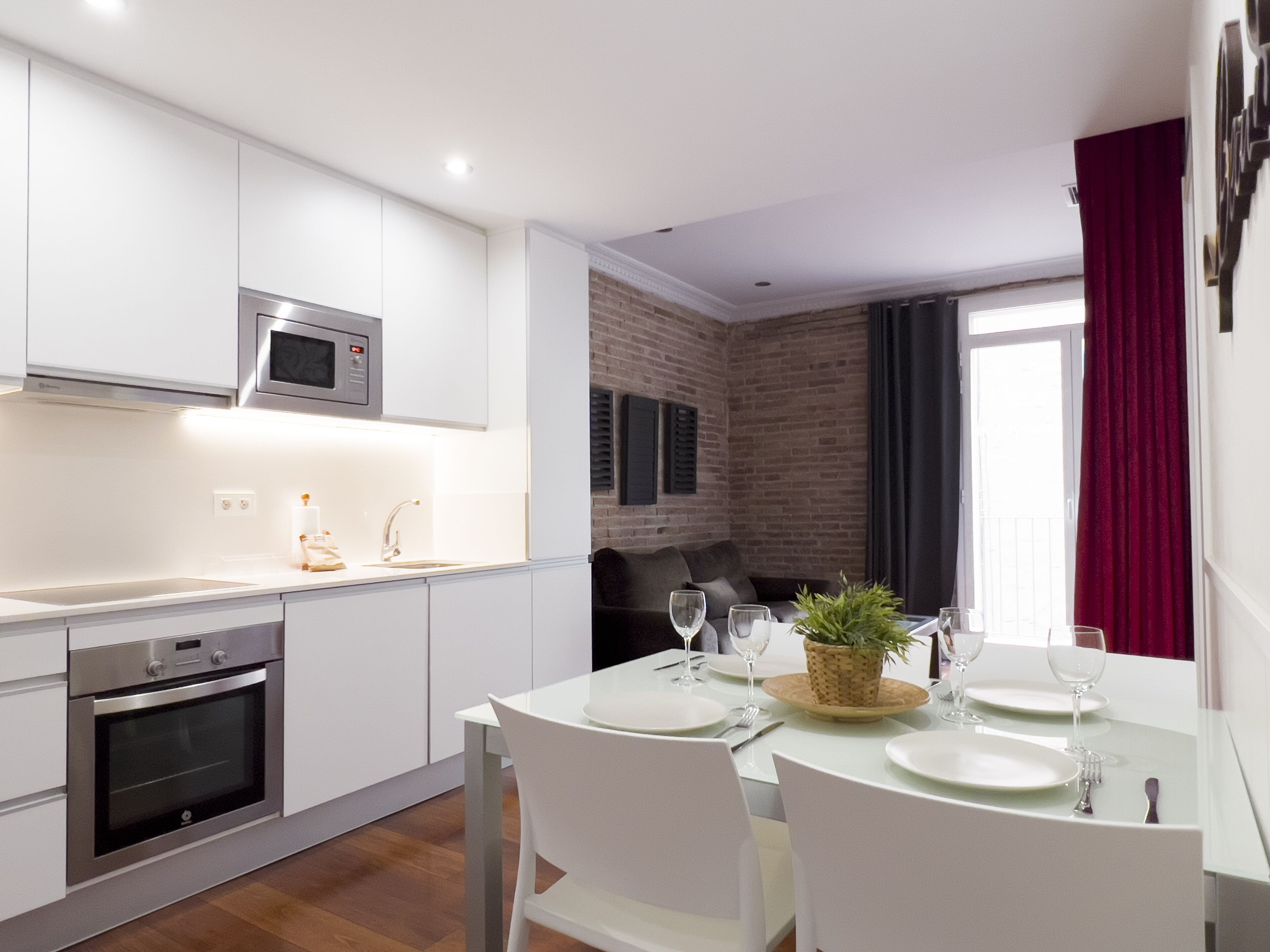 Una cocina moderna con armarios blancos, horno empotrado y microondas. Hay una zona de comedor con una mesa dispuesta para cuatro personas, incluyendo platos, vasos y cubiertos. Una planta en maceta está sobre la mesa, y se ve una pared de ladrillo al fondo.