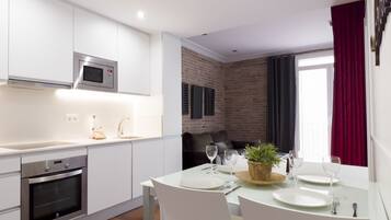 Una cocina moderna con armarios blancos, horno empotrado y microondas. Hay una zona de comedor con una mesa dispuesta para cuatro personas, incluyendo platos, vasos y cubiertos. Una planta en maceta está sobre la mesa, y se ve una pared de ladrillo al fondo.
