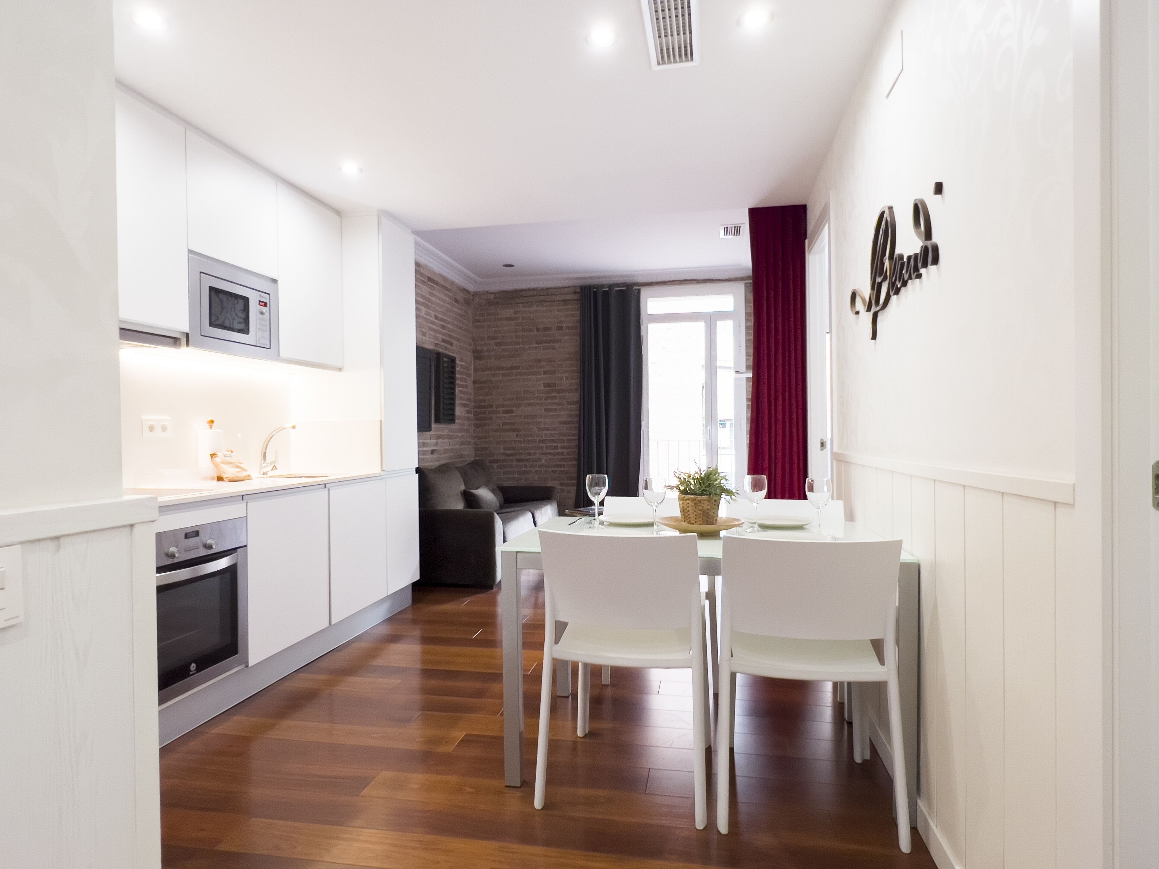 Una cocina moderna con armarios blancos, un microondas y un horno. Hay un comedor con una mesa y sillas, y una cortina roja.