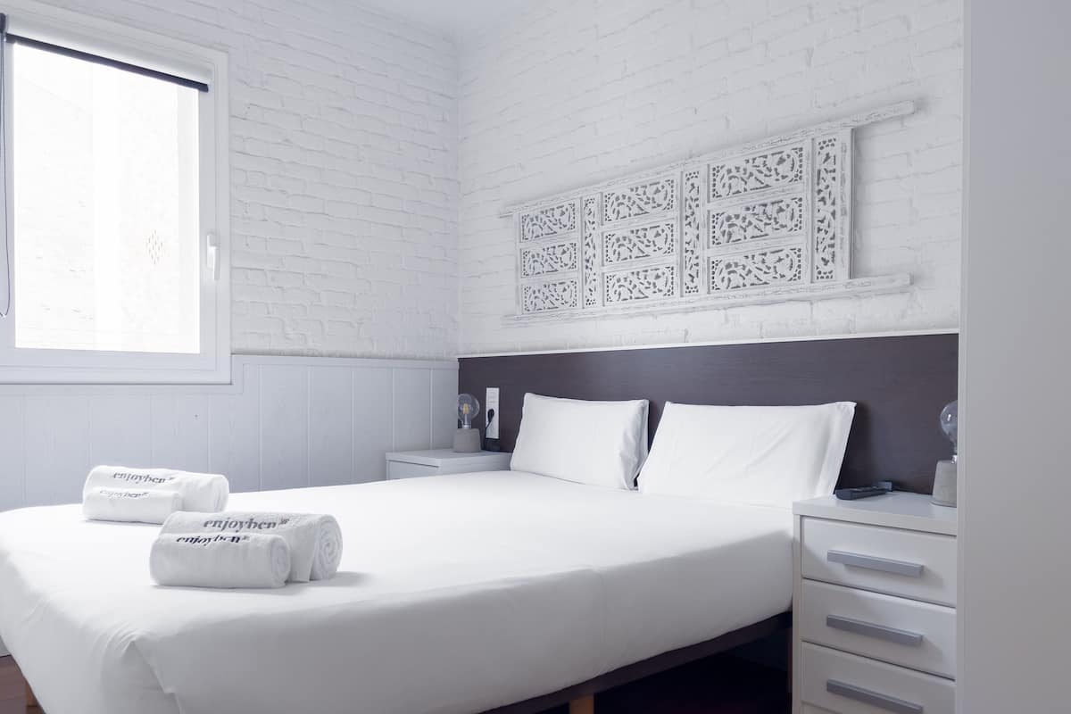 Un dormitorio moderno con una cama grande, ropa de cama blanca, una mesita de noche con lámpara y un panel decorativo en la pared.