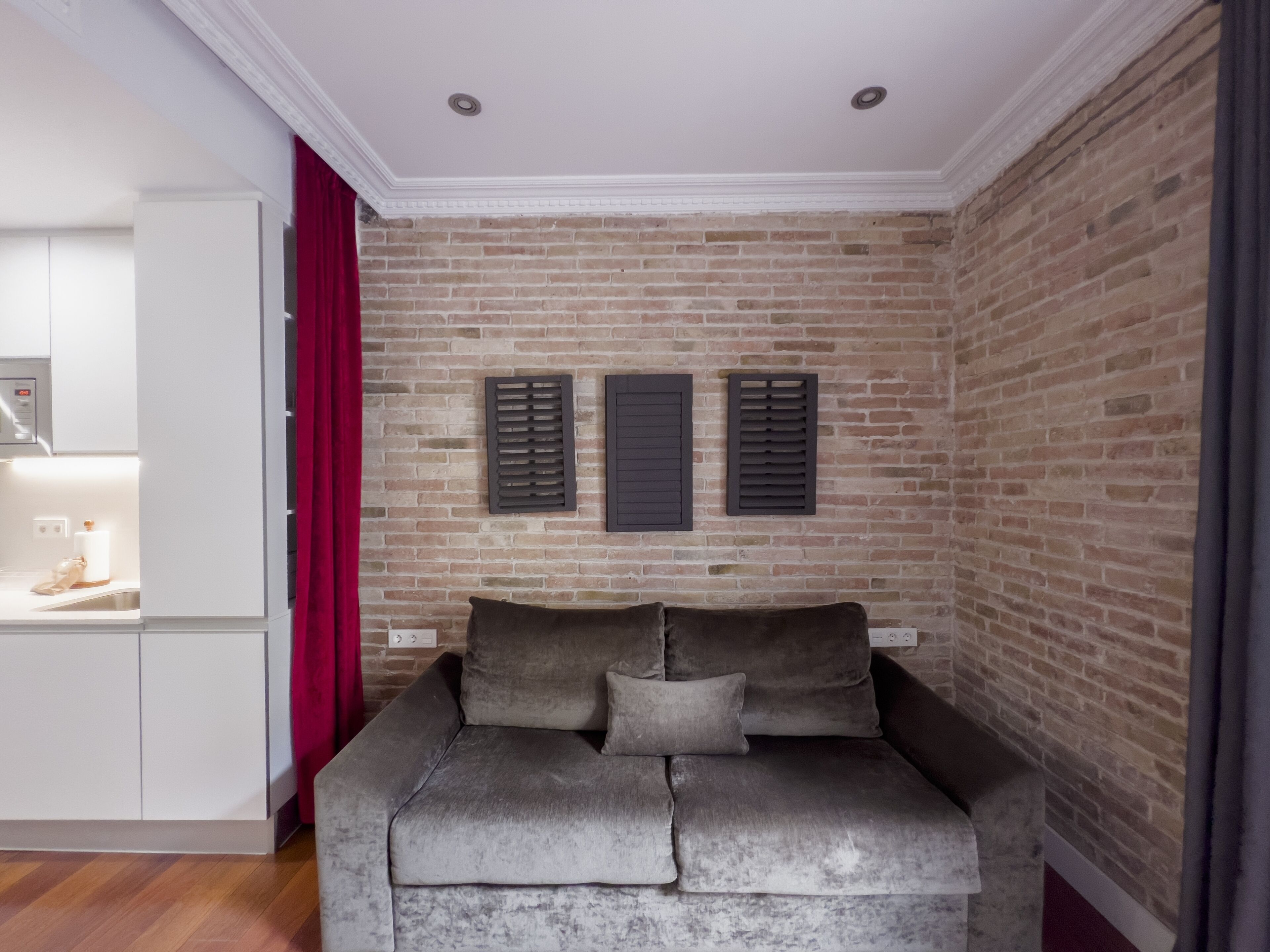 Una sala de estar con pared de ladrillo, un sofá con cojines, una cortina roja y una zona de cocina con armarios blancos.