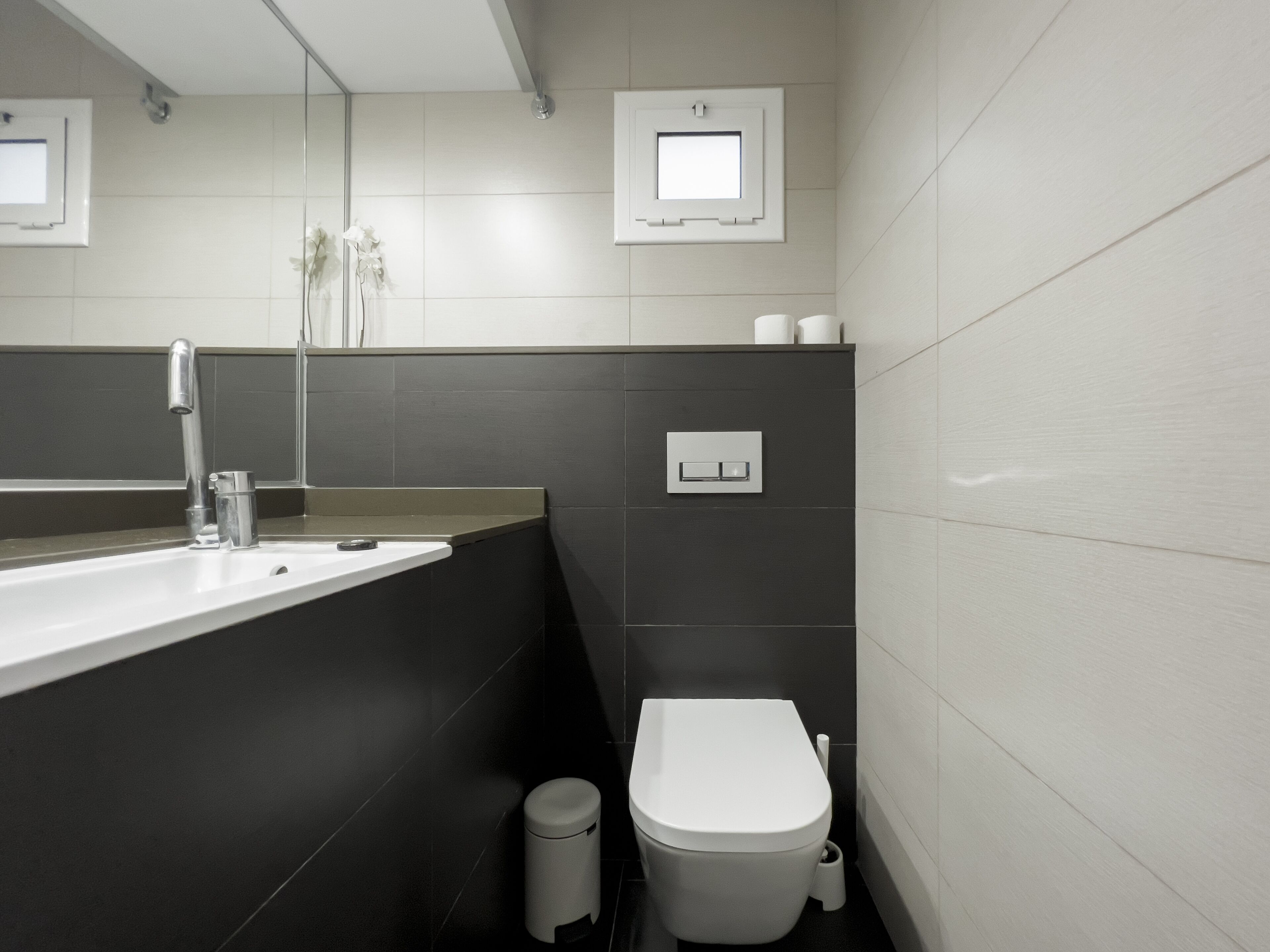 A modern bathroom with a white sink, dark tiles, a toilet, and a trash bin.