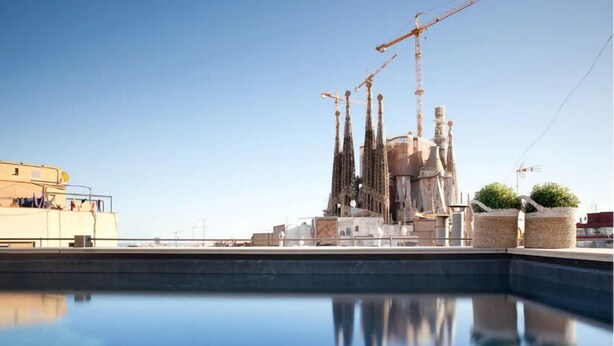 A pool reflecting a building with tall spires and construction cranes, surrounded by a clear sky. - Enjoybcn Gaudi Apartments (Barcelona)