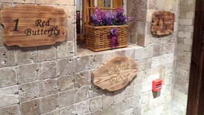 Un mur de pierre orné de panneaux en bois indiquant les numéros et les noms des pièces, un panier en osier rempli de fleurs violettes et un détecteur d’incendie.