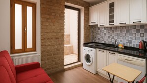 Un espace de vie compact comprenant un canapé rouge, un lave-linge, une petite table et un coin cuisine avec des meubles blancs et un pare-feu en carreaux.