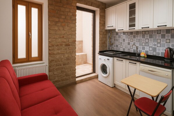 A compact living space with a red sofa, a washing machine, a small table, and a kitchen area with white cabinets and a tiled backsplash. - Istanberry - Pera Apartments (Istanbul)