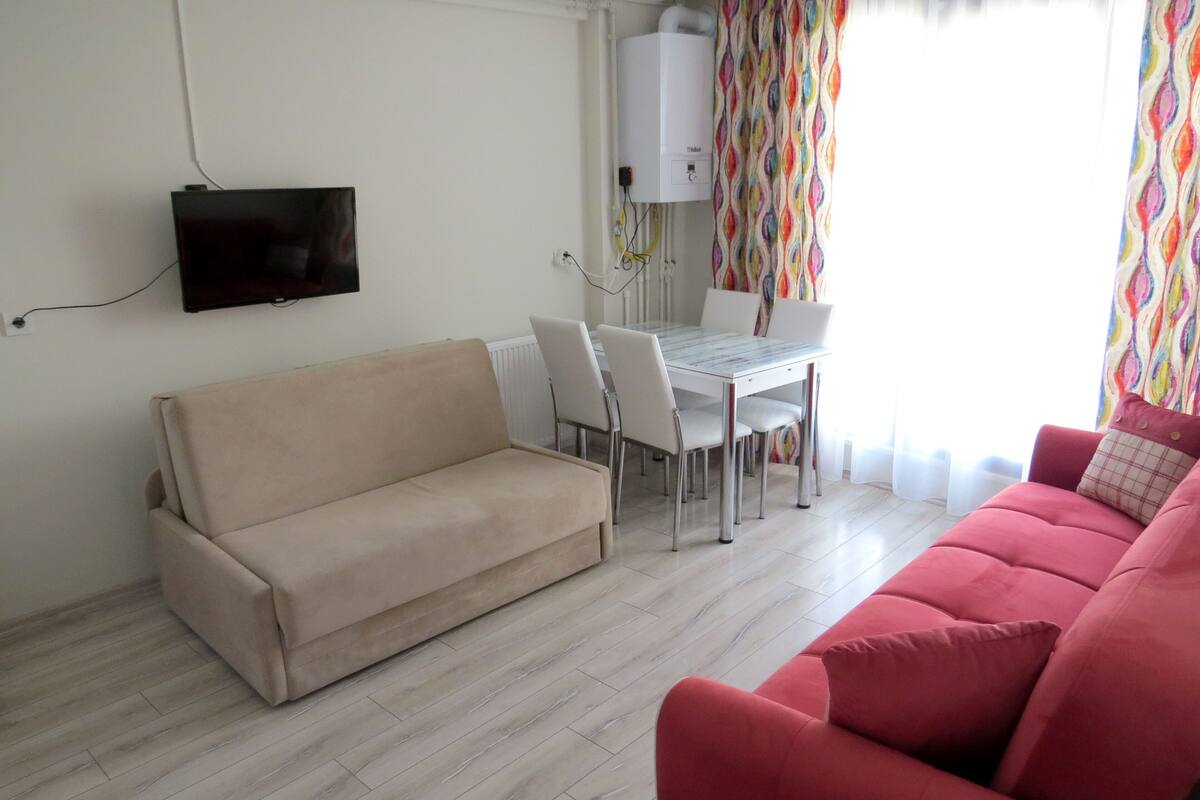 Una sala de estar con un sofá color beige, un sofá rojo, una mesa de comedor con sillas, un televisor montado en la pared y una cortina colorida.