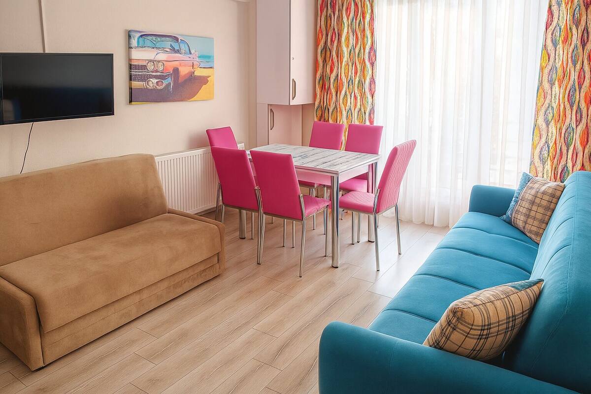 Una sala de estar con un sofá color arena, un sofá azul, una mesa de comedor con sillas rosadas, un televisor en la pared y una cortina colorida.