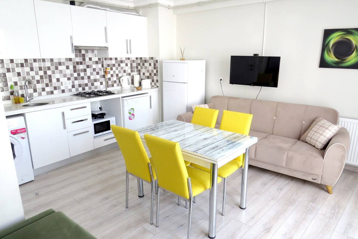 Una cocina moderna con armarios blancos, salpicadero de azulejos y un comedor con mesa y sillas amarillas.