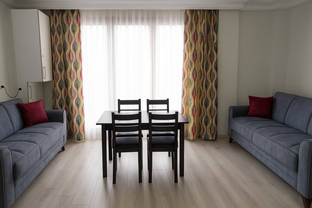Una sala de estar con dos sofás grises, una mesa de comedor con cuatro sillas y cortinas coloridas.