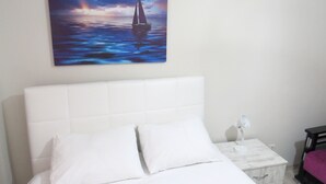 A neatly made bed with white bedding and pillows, a white headboard, a small bedside table with a lamp, and a painting of a sailboat on the wall. - Istanberry - Paradise Apartments (Istanbul)