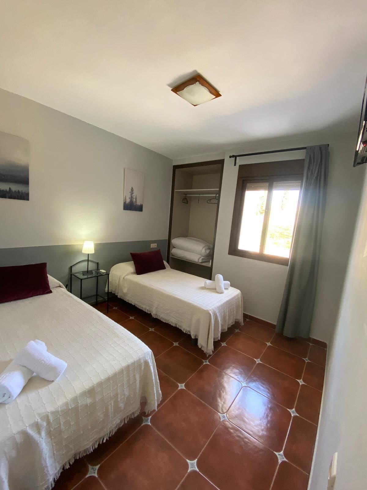 A hotel room with two beds, a wardrobe, a window with curtains, and a nightstand with a lamp.
