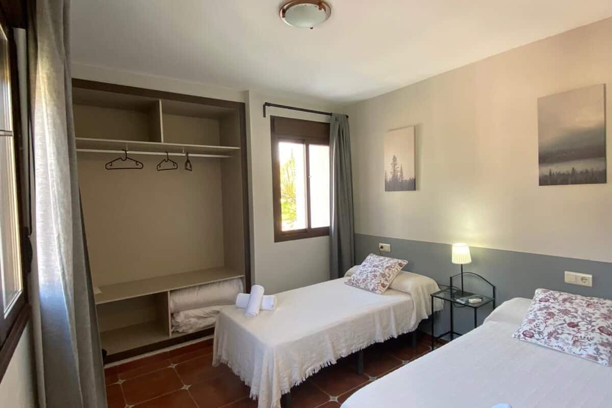 Une chambre d’hôtel avec deux lits, une armoire, une fenêtre avec des rideaux et une petite table avec une lampe.