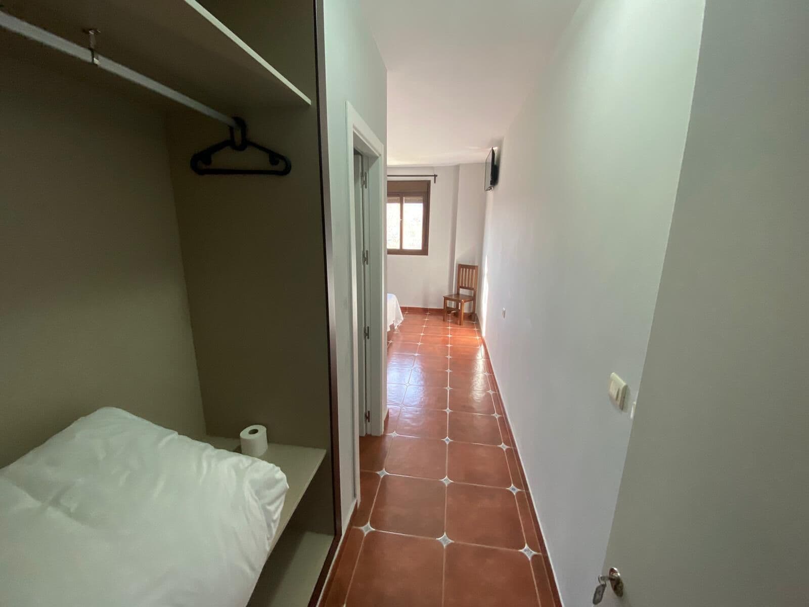 A narrow hallway with a bed on the left, a chair in the middle, and a door at the end.