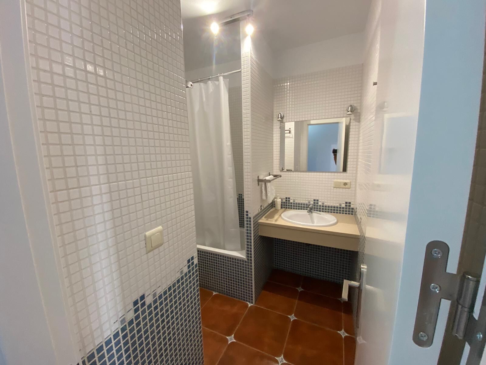 A bathroom with white tiled walls, a shower, a sink with a mirror, and a door with a handle.