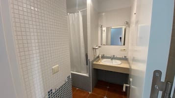 A bathroom with white tiled walls, a shower, a sink with a mirror, and a door with a handle.
