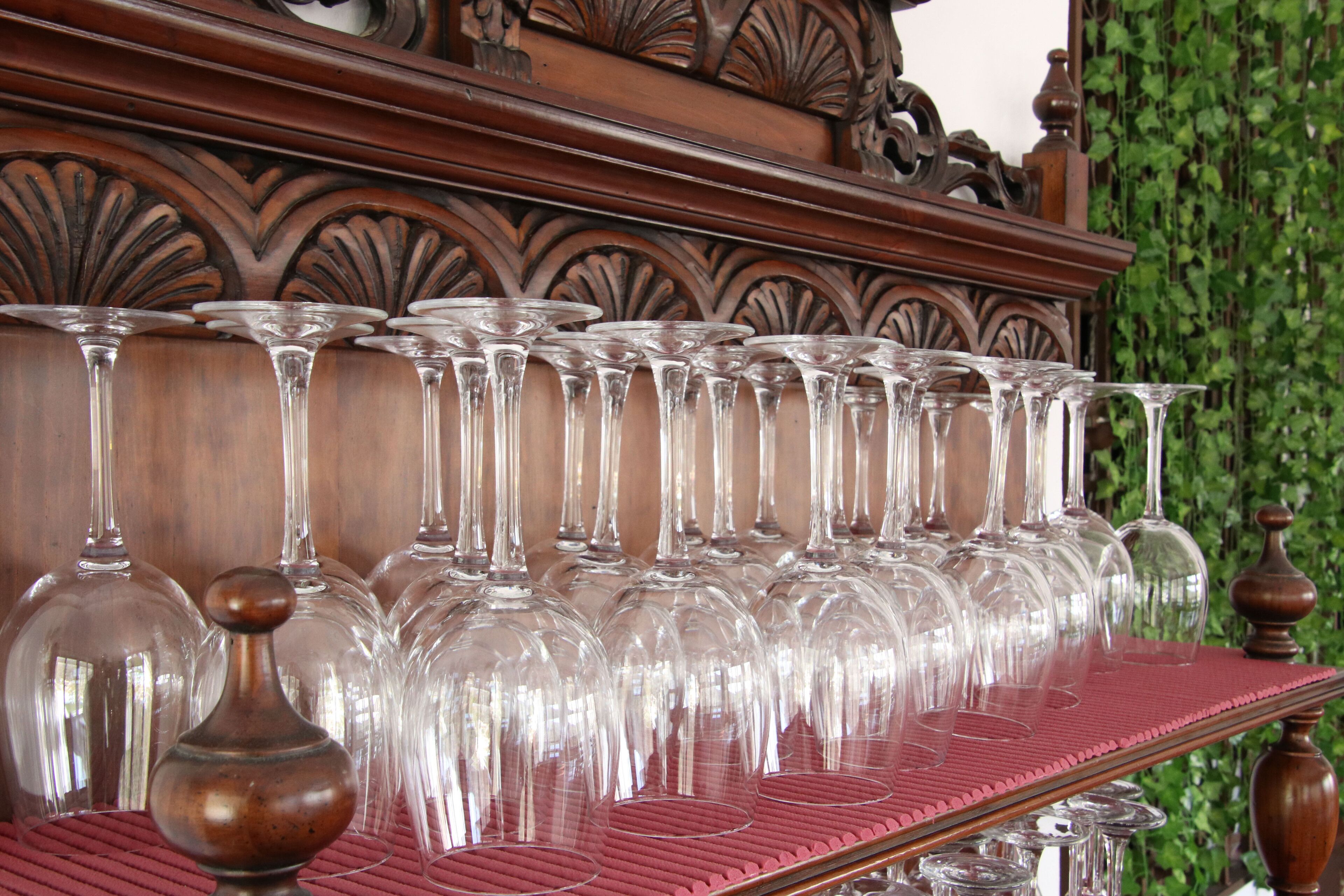 Un hilera de copas de cristal transparente sobre una superficie de madera con un tapete rojo, frente a un cabecero de madera tallada y follaje verde.