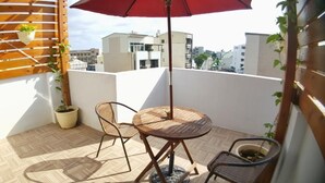 Ein kleiner Balkon mit runder Holztisch und zwei Stühlen, einem roten Sonnenschirm und Blumentöpfen.