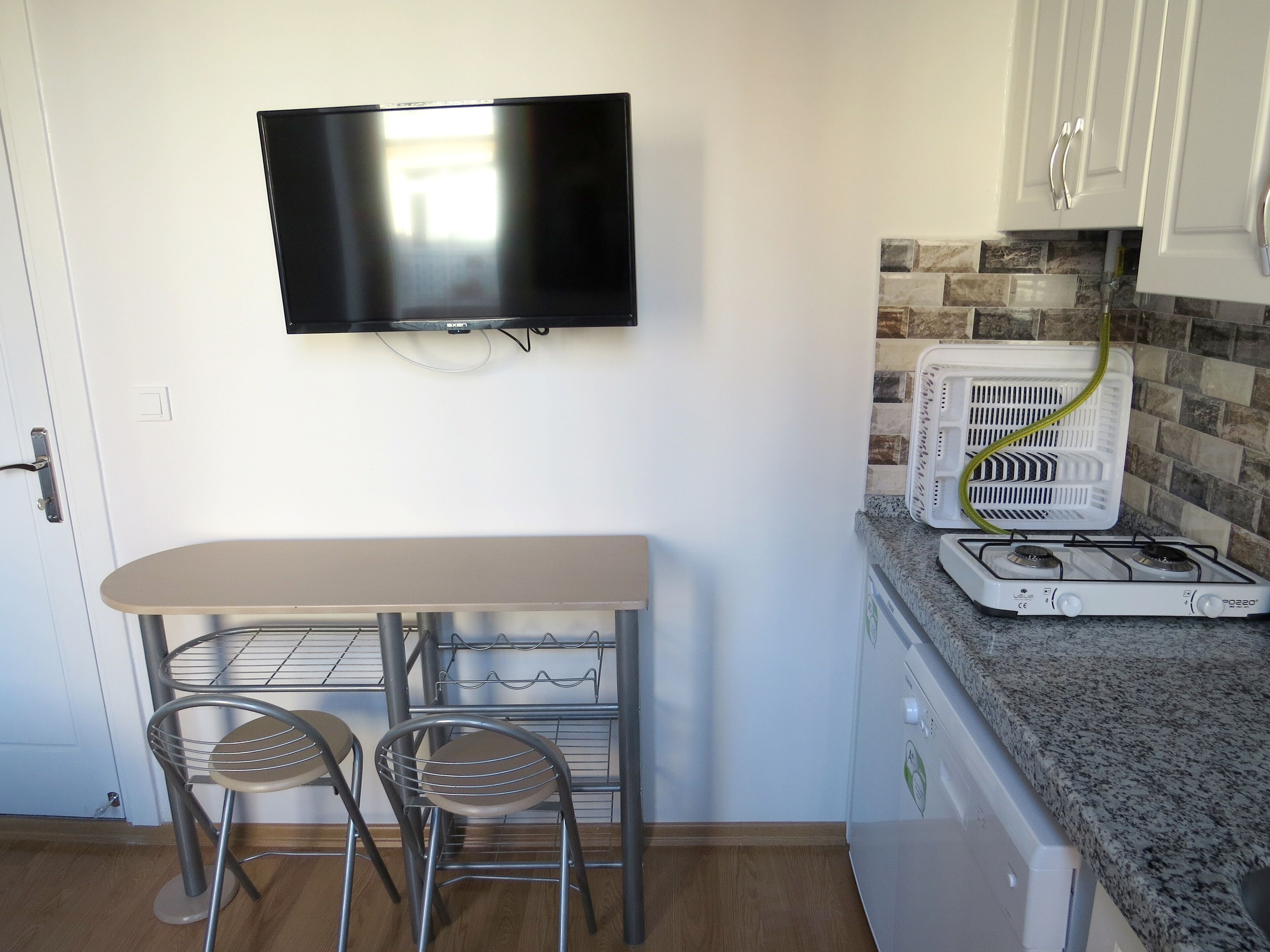 Cocina compacta con un televisor en la pared, una pequeña mesa de comedor con dos sillas y una estufa a gas.
