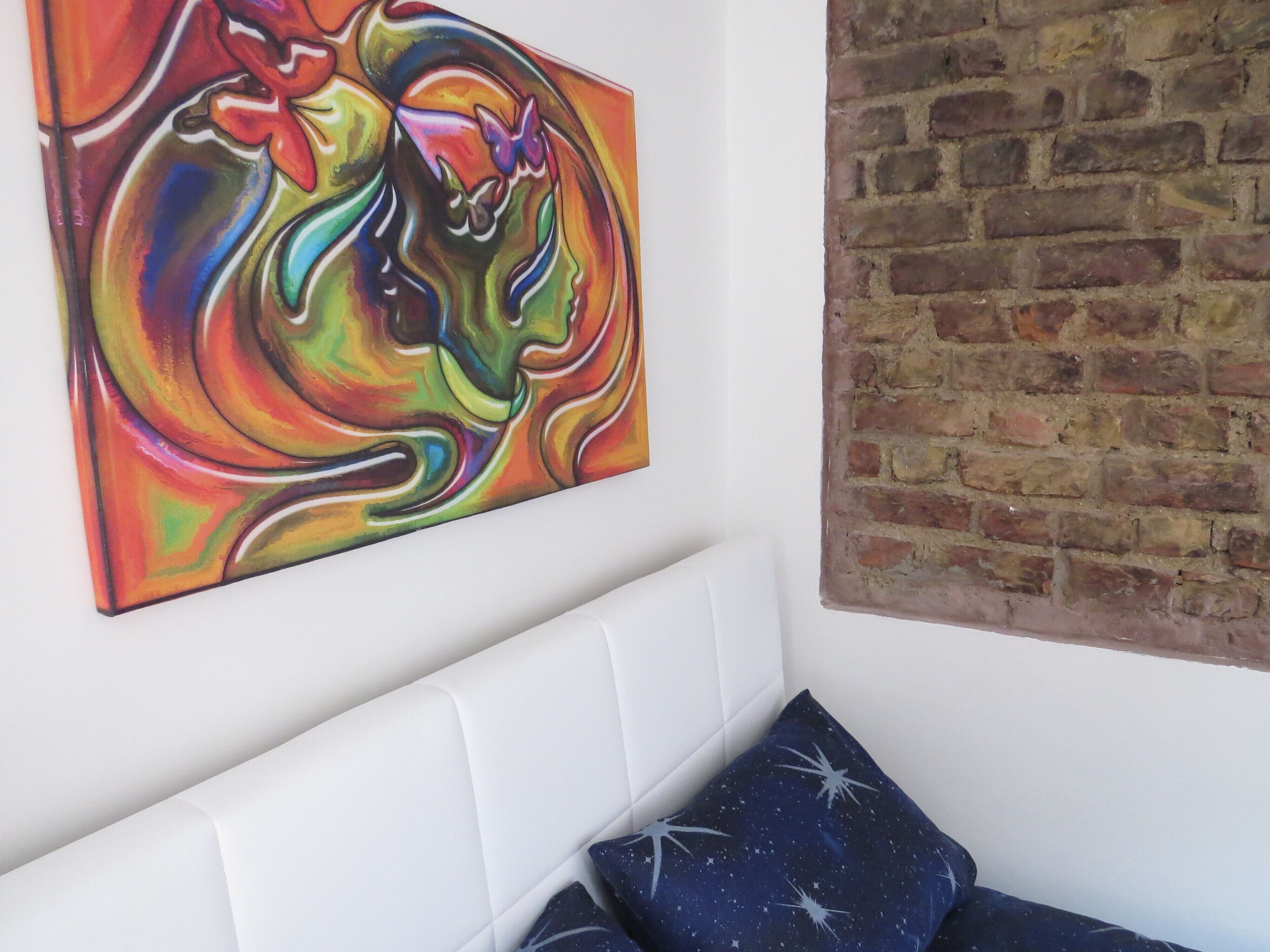 Una habitación con un cabecero blanco, un cuadro abstracto colorido y una pared de ladrillo.