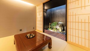 Une pièce de style japonais traditionnel, avec un sol recouvert de tatamis, une table en bois agrémentée d’un plateau décoratif et une porte coulissante donnant sur un jardin où se trouve une lanterne en pierre.