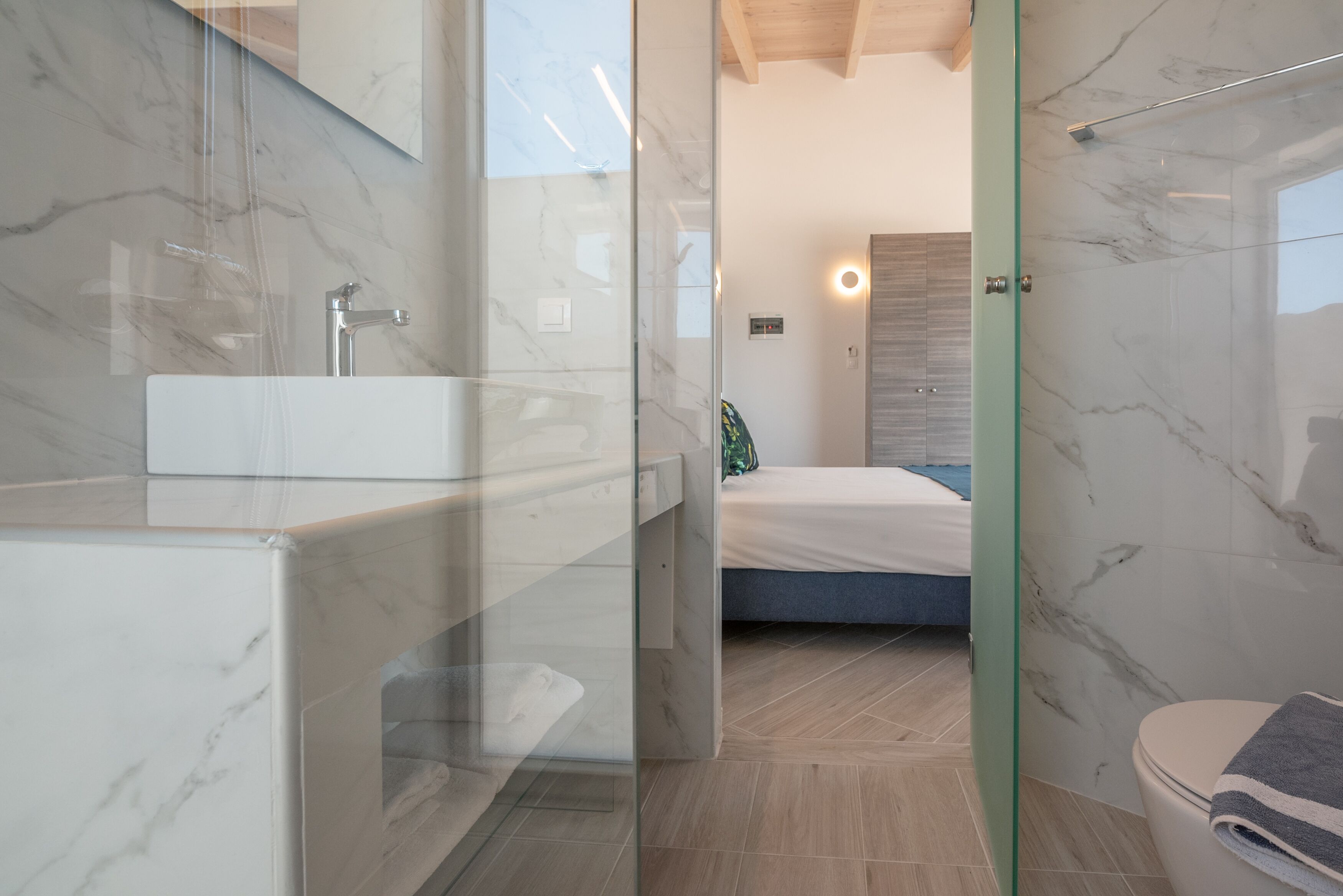 A modern bathroom with a glass-enclosed shower, a marble sink, and a visible bed in the background.