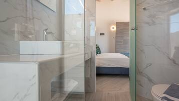 Un baño moderno con ducha de vidrio, lavamanos de mármol y una cama visible al fondo.