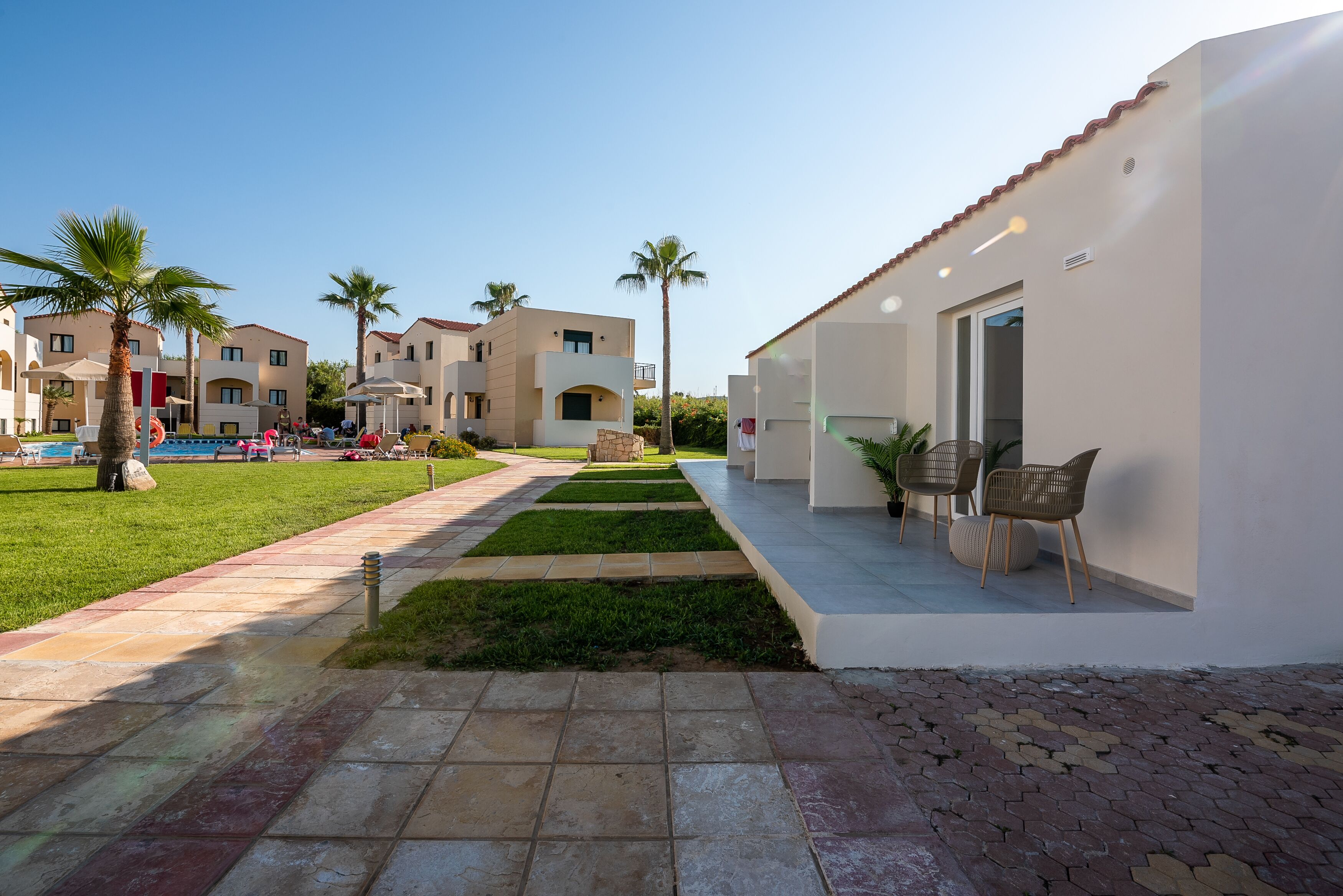 Un resort con zona de piscina, palmeras y un camino que lleva a un edificio con asientos al aire libre.