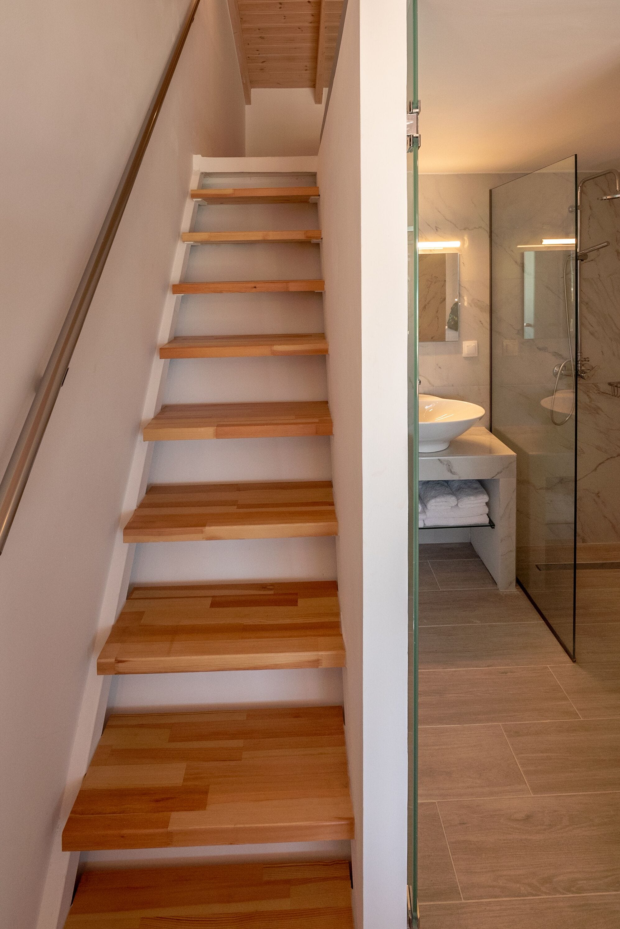 Een moderne trap met houten treden die leidt naar een badkamer met een douchecabine van glas en een marmeren wastafel.